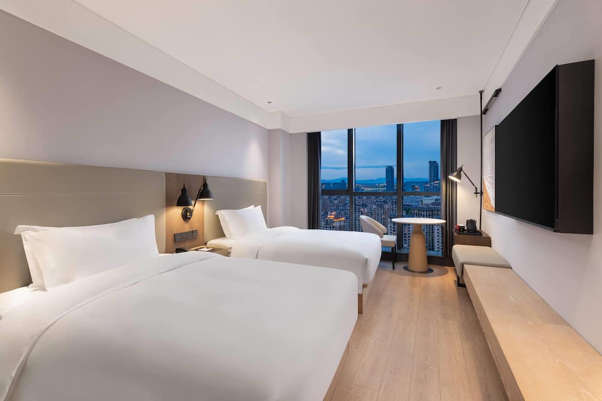 Habitación de hotel moderna con dos camas, un escritorio, una silla y un amplio ventanal con vista a la ciudad.