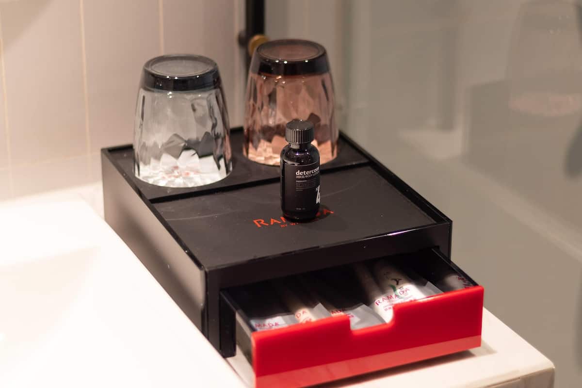 Una encimera de baño con una bandeja negra que contiene una botella de detergente, un recipiente de vidrio y un cajón rojo con artículos de aseo.