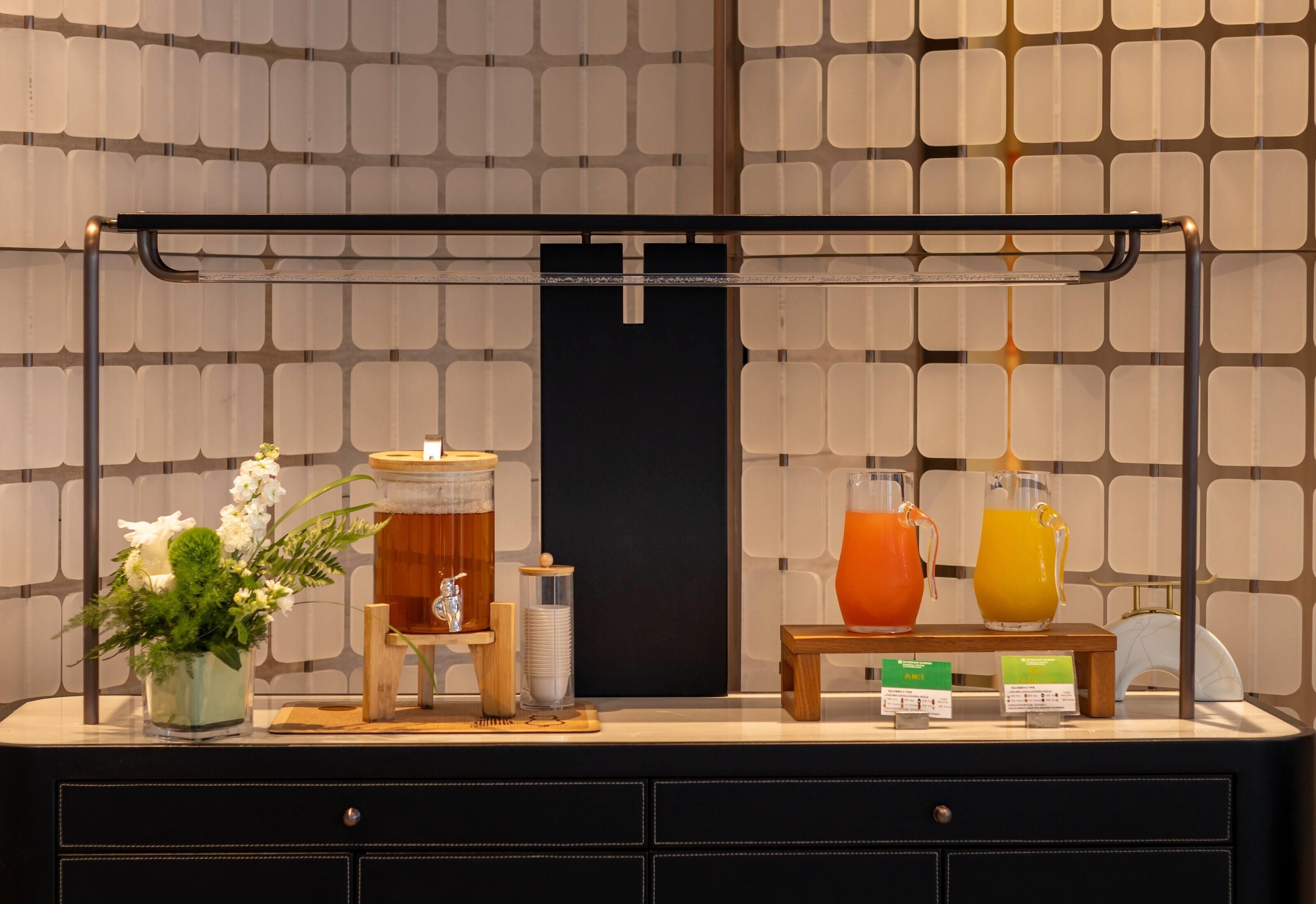 Een bar met een drankstation, inclusief een glas sinaasappelsap, een glas ijsthee en een karaf sinaasappelsap. Er staat een klein houten tafeltje met een bloevas en een glas water.