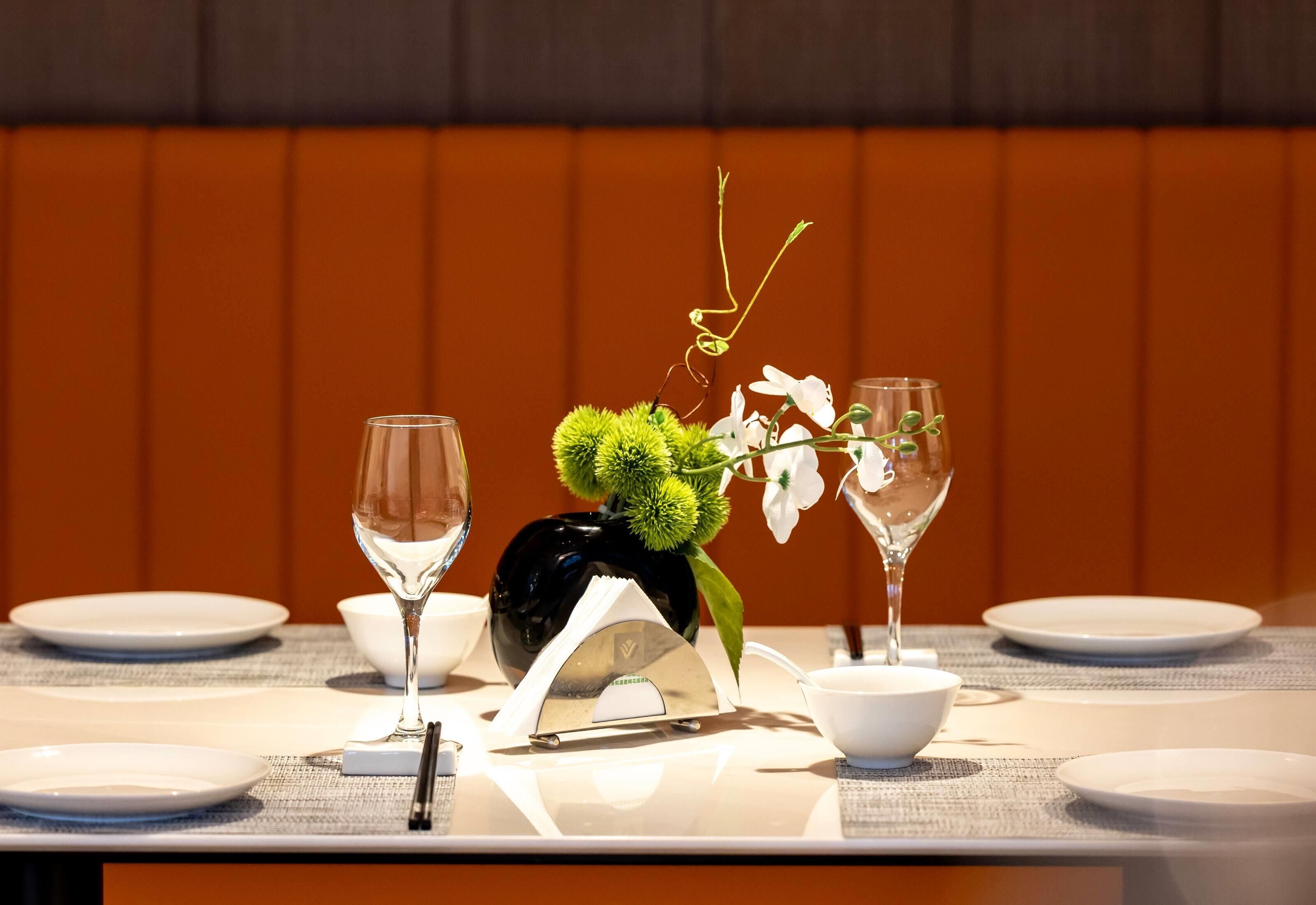 Une table à manger dressée avec de l’assiette, des bols et des couverts blancs, agrémentée d’un vase contenant des feuilles vertes et des fleurs blanches.