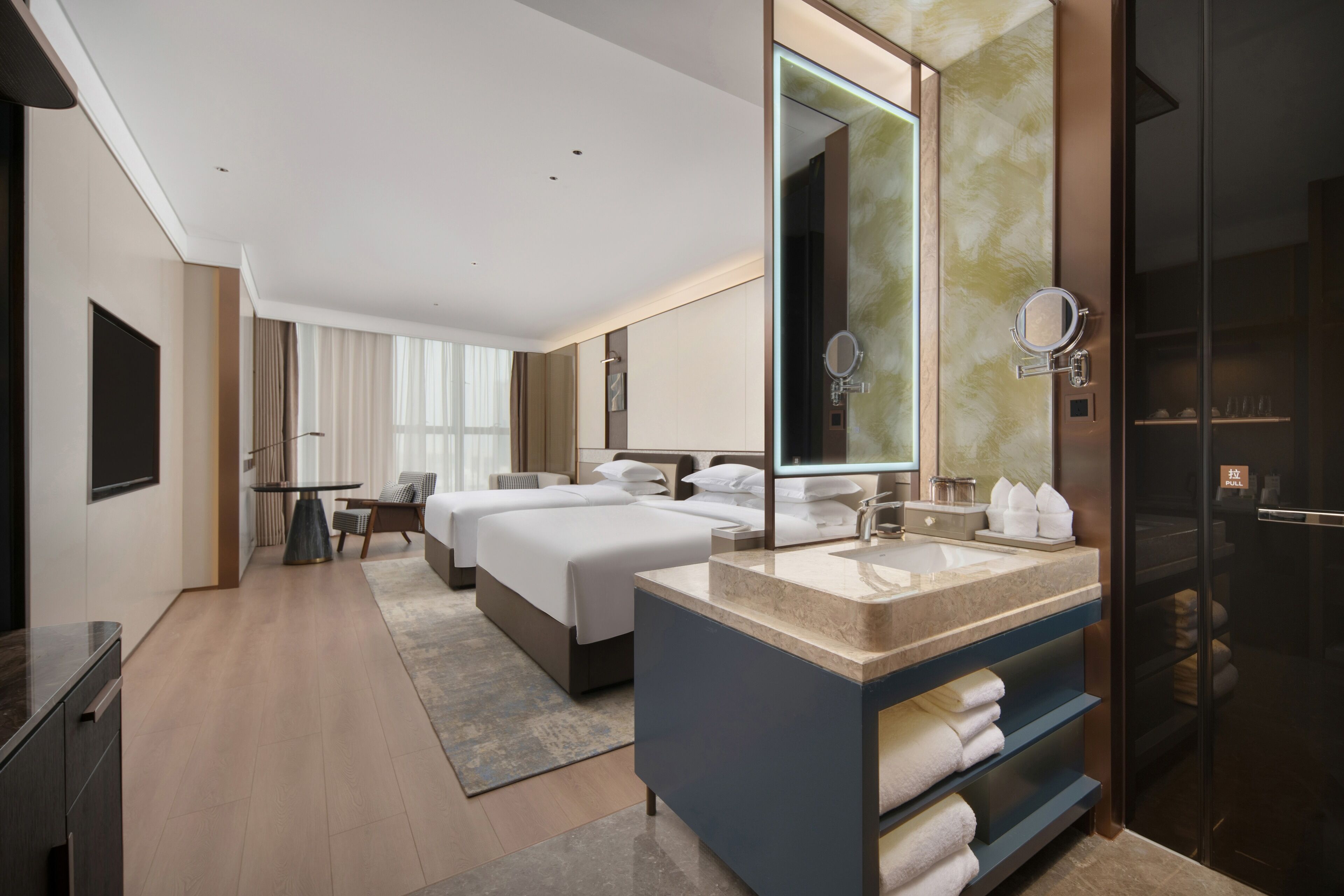 A hotel room with a large bed, a separate bed, a bathroom with a mirror and towel rack, and a television.