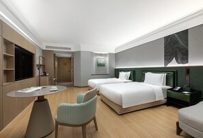 Quarto de hotel com cama, duas mesas de cabeceira, uma cadeira, uma mesinha, televisão e um quadro na parede.