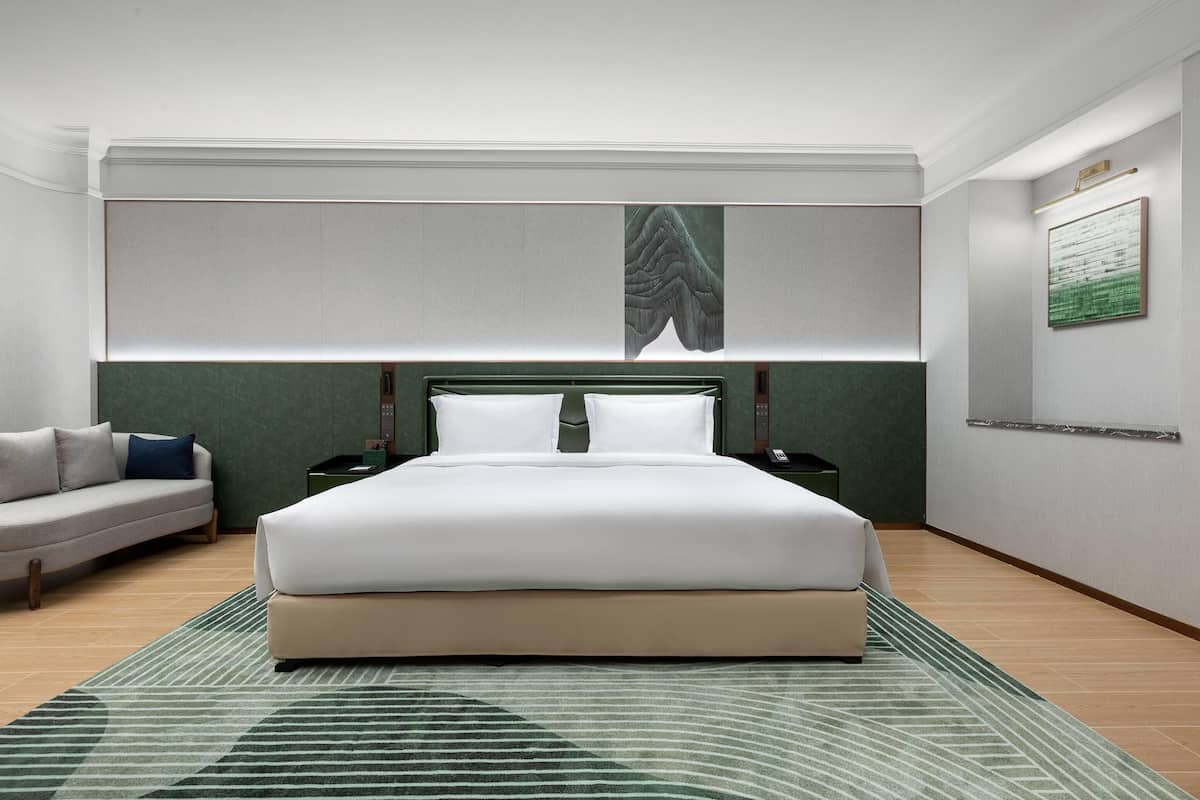 A modern hotel room with a large bed, a sofa, a nightstand, and a wall-mounted artwork.
