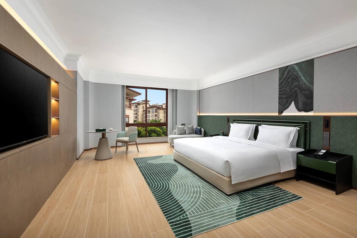 A modern hotel room with a large bed, a flat-screen TV, a small table with chairs, and a view of buildings through the window.