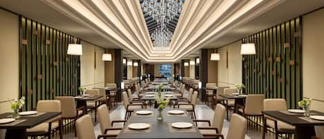 A spacious hotel dining area with long tables, chairs, and a central chandelier.