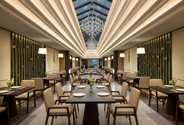 Une vaste salle à manger d’hôtel, dotée de longues tables, de chaises et d’un lustre central.
