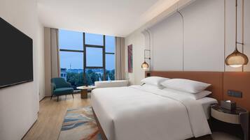 A modern hotel room with a large bed, a desk, a chair, and a view of the cityscape through large windows.