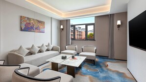Una habitación de hotel moderna con sofá, sillas, una mesa de centro, un cuadro, televisor y un ventanal con vistas a la ciudad.