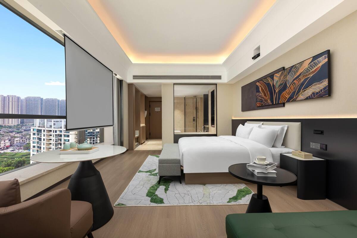 A modern hotel room with a large bed, a desk, a chair, a small table, and a large window offering a city view.