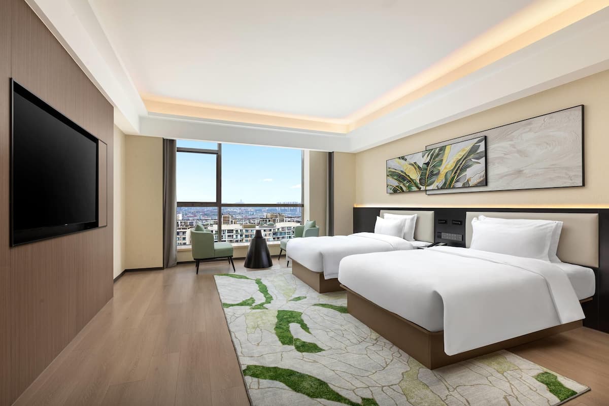 A modern hotel room with two beds, a flat-screen TV, a city view, and a large window.