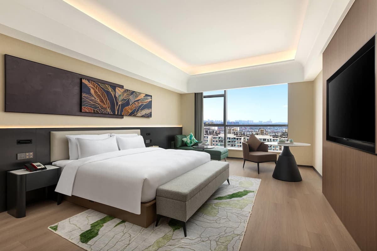 A modern hotel room with a large bed, a seating area, a TV, and a city view.