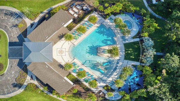 An aerial view of a resort with a central swimming pool, surrounded by lounge areas and palm trees.