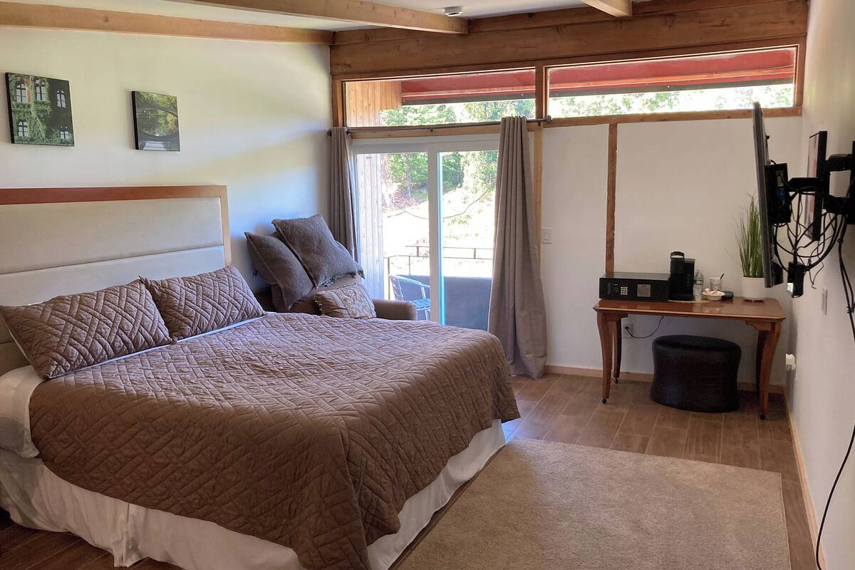 A bedroom with a large bed, a wooden headboard, a small table with a plant, and a window with curtains.