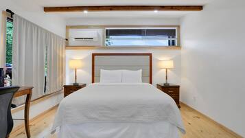 A bedroom with a large bed, two bedside tables with lamps, a window with blinds, an air conditioning unit, and a wooden floor.
