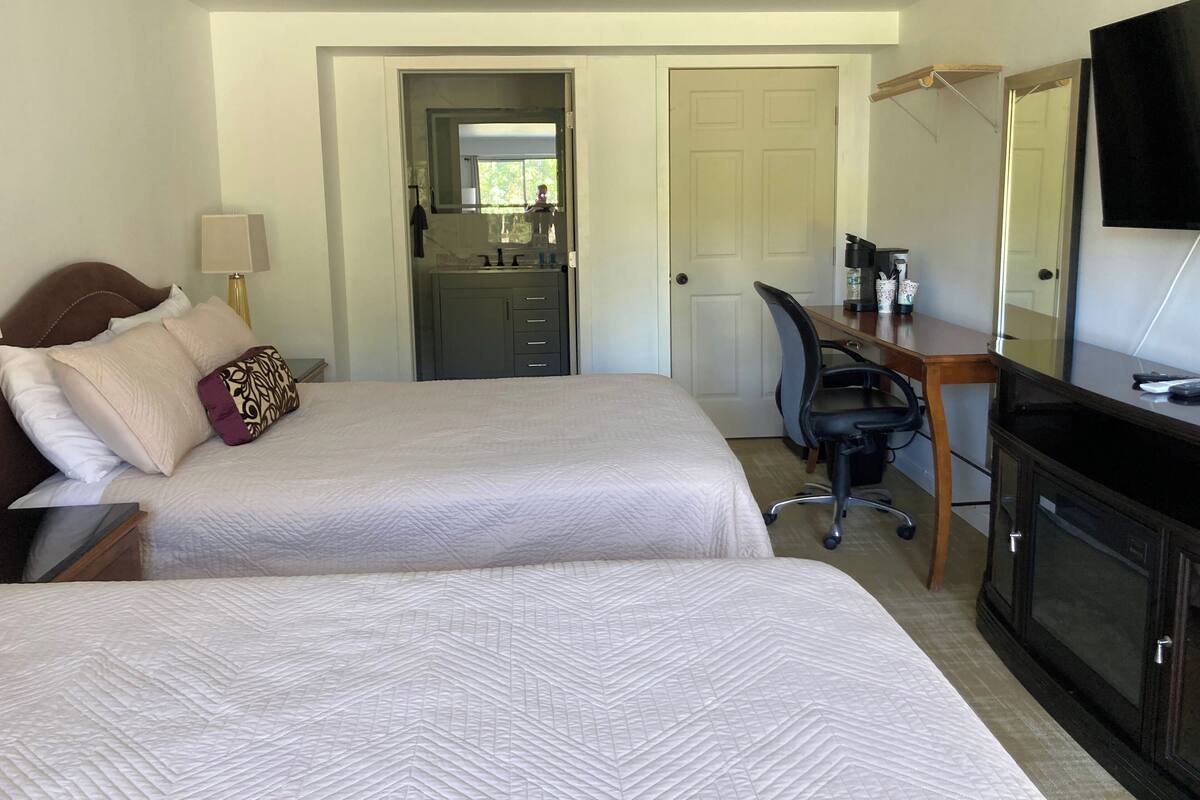 A hotel room with two beds, a desk, a chair, a television, and a bathroom visible through an open door.