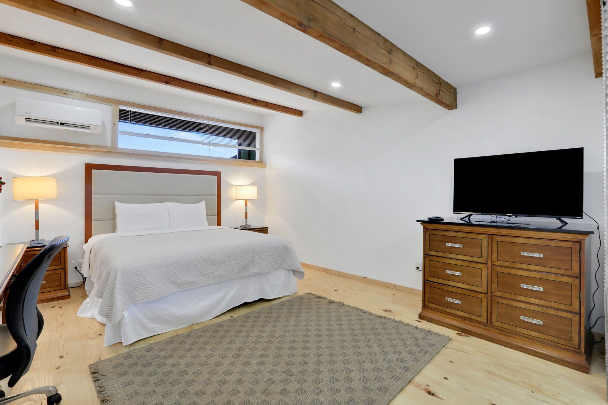 Ein Schlafzimmer mit einem Bett, Nachttischlampen, einem Fernseher auf einer Holzkommode und einem Schreibtisch mit Stuhl.