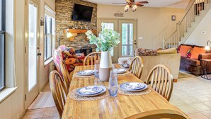 A dining area with a wooden table set for six, surrounded by chairs. A vase with flowers is on the table, and a television is mounted on the wall. - Pickwick Lake Cabin w/ Private Marina Access (Iuka)