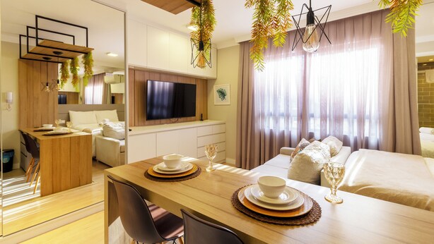 Una habitación de hotel moderna con una mesa de comedor de madera, una cama con ropa de cama color beige, un televisor montado en la pared y un ventanal con cortinas.