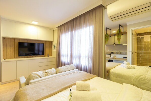 A hotel room with a large bed, a TV mounted on the wall, a bathroom with a shower, and a small kitchen area. - Studio Moderno Ap 67 (Curitiba)