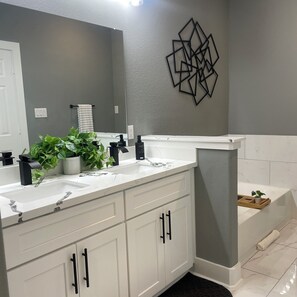 Une salle de bain avec un meuble-lavabo blanc, un grand miroir, un motif géométrique sur le mur et une baignoire dotée d’un plateau en bois.
