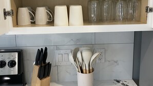 Et køkken med et træskab fyldt med hvide skåle, målebægre og glaskrus. Under skabet er en bordplade med et knivblok og en grydeholder.