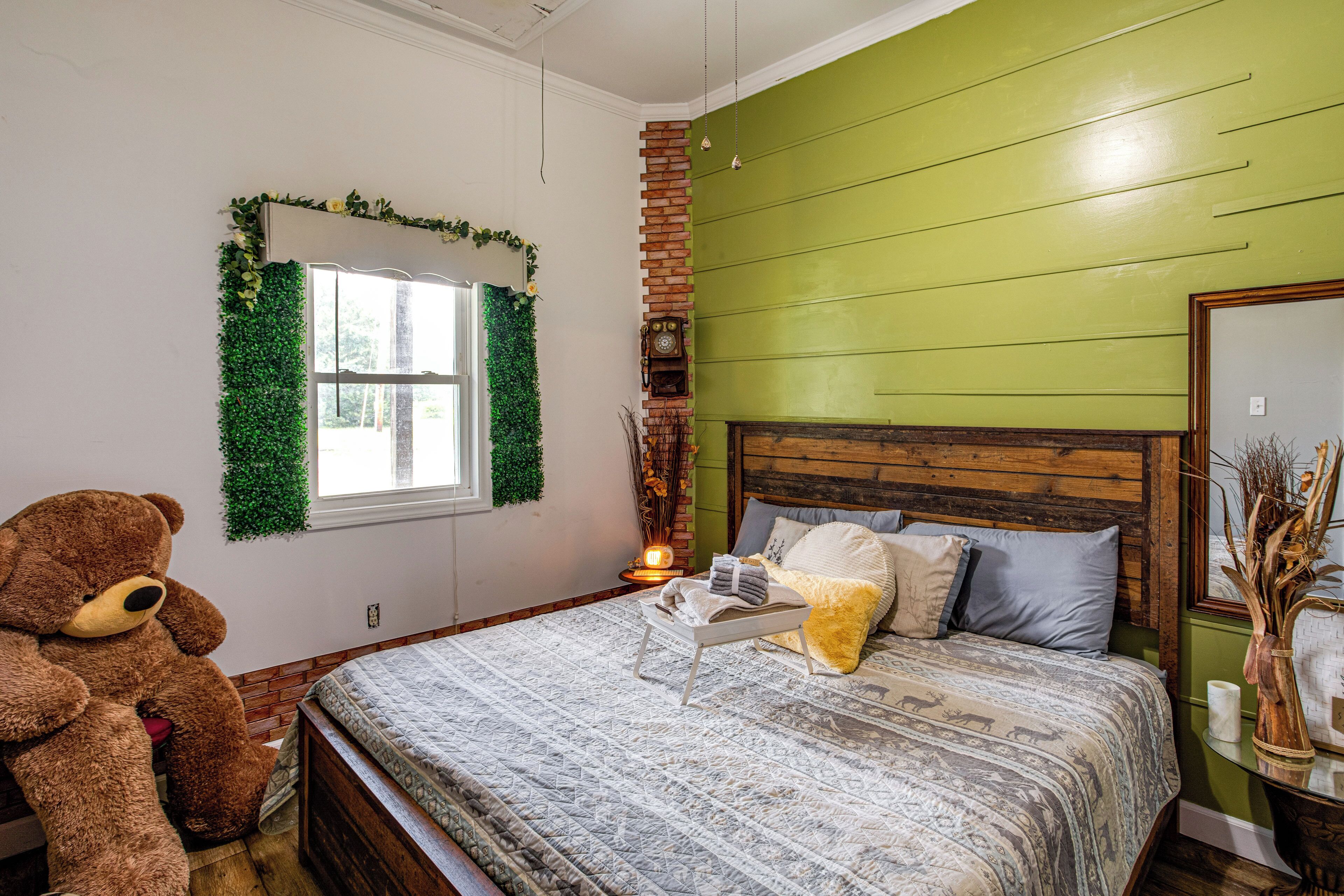 A bedroom with a large bed, a teddy bear, a window with green foliage, and a brick wall.