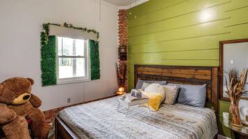 A bedroom with a large bed, a teddy bear, a window with green foliage, and a brick wall.