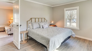 A bedroom with a bed, a nightstand, a lamp, a window, and a door leading to another room.