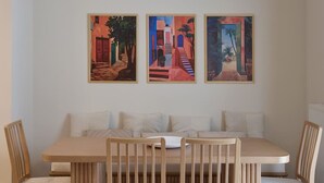 A dining area with a wooden table and chairs, a bench with cushions, and three colorful paintings on the wall. - Prestige Lifestyle 3bd Apt in Kolonaki (Athina)