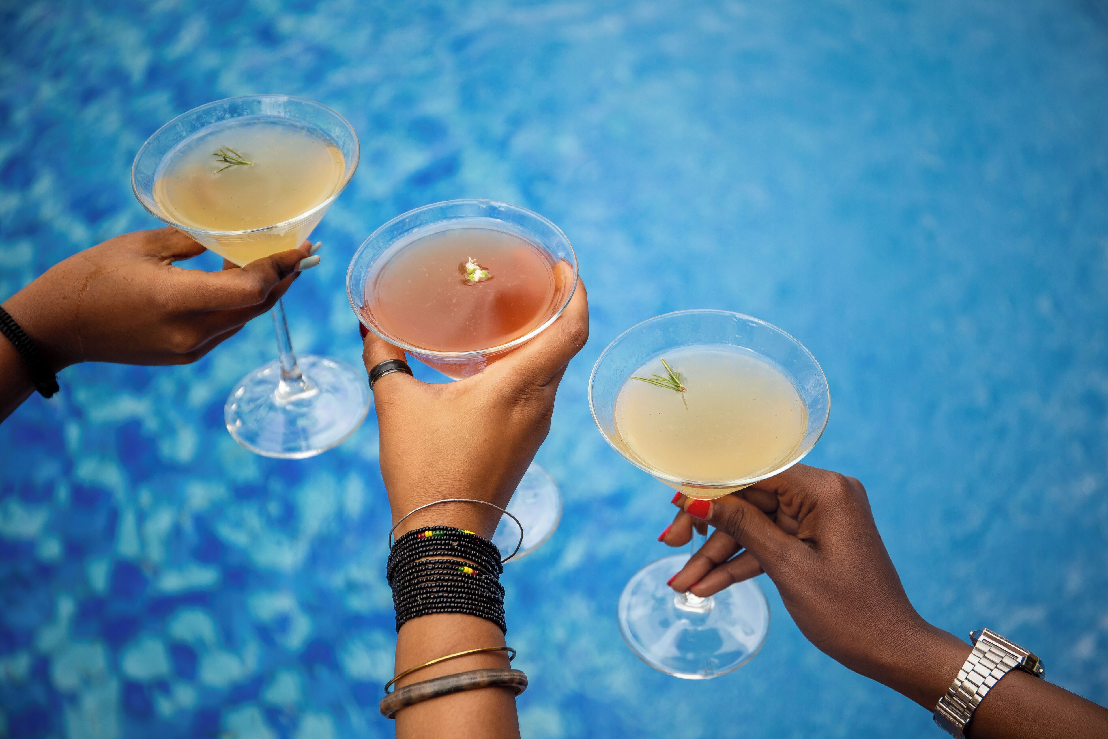 Tre mani che sorreggono bicchieri da martini con un liquido trasparente e una guarnizione, su sfondo blu.
