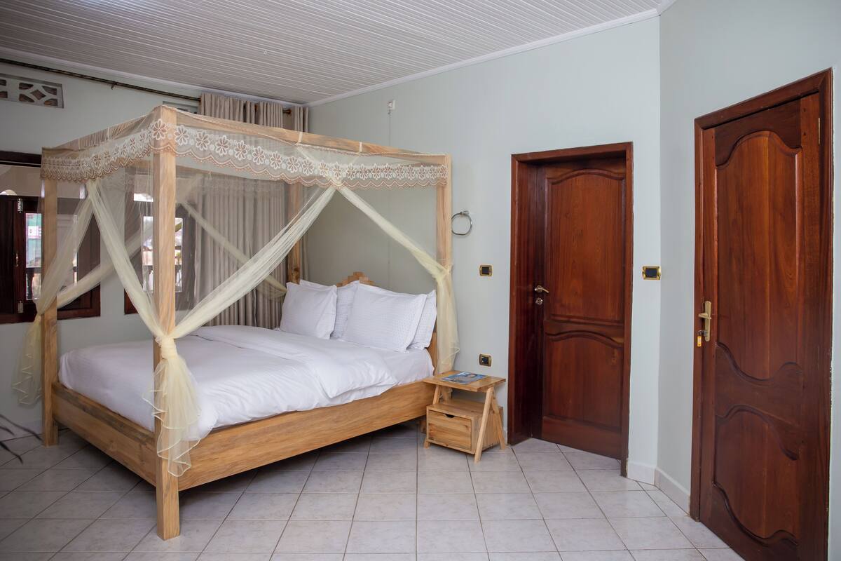 A four-poster bed with a wooden frame and white bedding, a small wooden nightstand, and a wooden door with a brass handle.