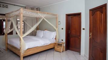 Un lit à baldaquin avec une structure en bois et du linge de lit blanc, une petite table de chevet en bois et une porte en bois dotée d’une poignée en laiton.