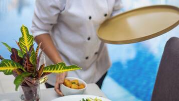 A chef serving food by a poolside table with a plant, a bowl of fruit, and a glass of white wine.