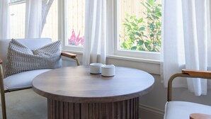 A room with a round wooden table, white curtains, a sofa, and a view of greenery outside. - Walk to Downtown - Luxe Backyard - Private + Peaceful Stay (St. Augustine)