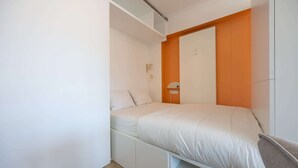 Une petite chambre à coucher moderne, avec un lit, des murs blancs, un mur d’accent orange et des rangements intégrés.