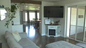 En moderne stue med sofa, TV og pejs. I baggrunden ses et spiseområde, og en skydedør fører til en udendørs terrasse.
