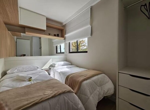 A modern bedroom with two beds, a built-in wardrobe, and a window with a view of trees. - Studio Moderno Ap 21 (Curitiba)
