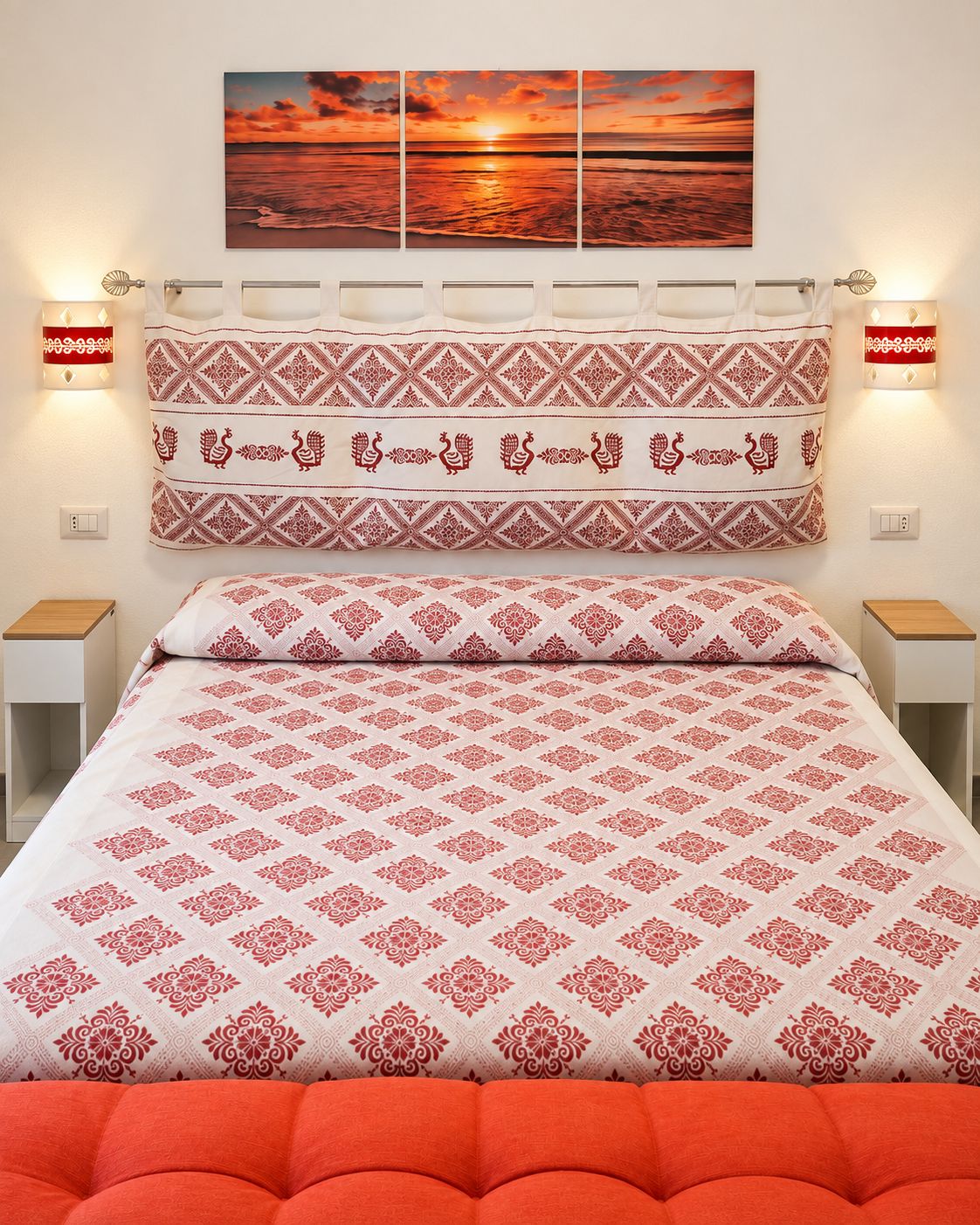 A bedroom with a patterned bedspread, a decorative headboard, and a sunset painting above the bed.