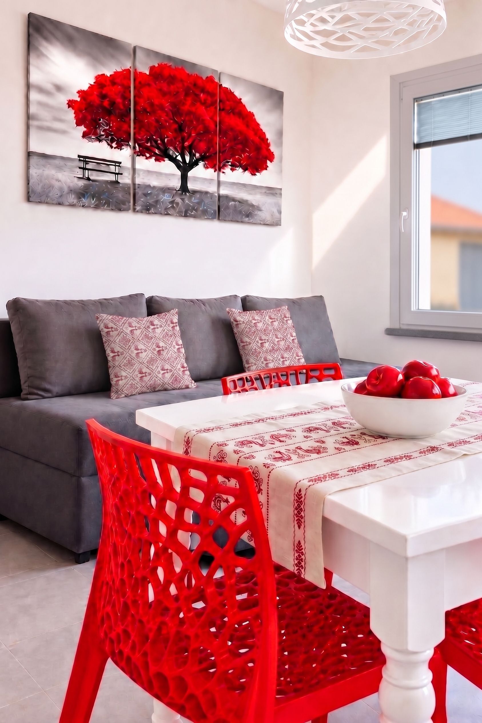 A modern living room with a grey sofa, red patterned cushions, a white table with a red bowl, and a red chair. A large red tree painting is on the wall.