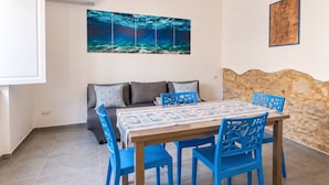A dining area with a table set for four, blue chairs, a sofa with cushions, a wall-mounted air conditioner, and a large ocean-themed painting. - Room-private Blue Apartment at Palma de (Sant'Antioco)