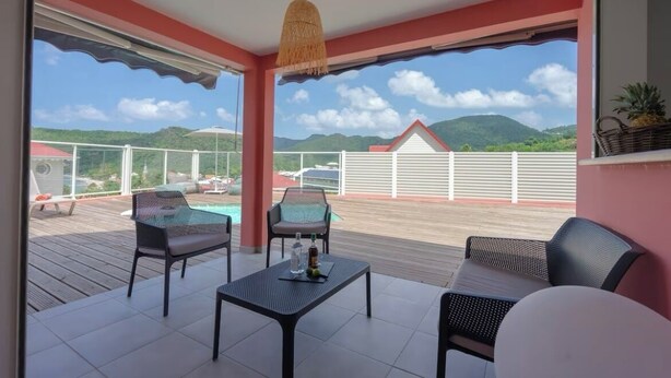 Un espace extérieur aménagé, abrité et doté de sièges, avec vue sur la piscine, comprenant une table et des chaises, un luminaire suspendu, ainsi qu’une vue sur les montagnes et un bâtiment au toit rouge.