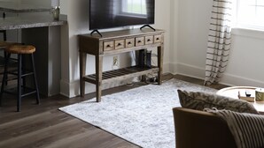 Een woonkamer met een televisie op een houten standaard, een barstoel, een kamerkleed met patroon en een raam met gordijnen.