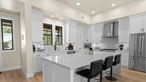 Une cuisine moderne avec des meubles blancs, un grand îlot central doté de tabourets, des appareils électroménagers en acier inoxydable et une vue sur l’extérieur.