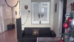 Ein Wohnzimmer mit einer schwarzen Couch, einem kleinen Tisch mit Zeitschriften, einem Weihnachtsbaum mit Dekorationen und einem Fenster mit einem dekorativen Gegenstand darüber.