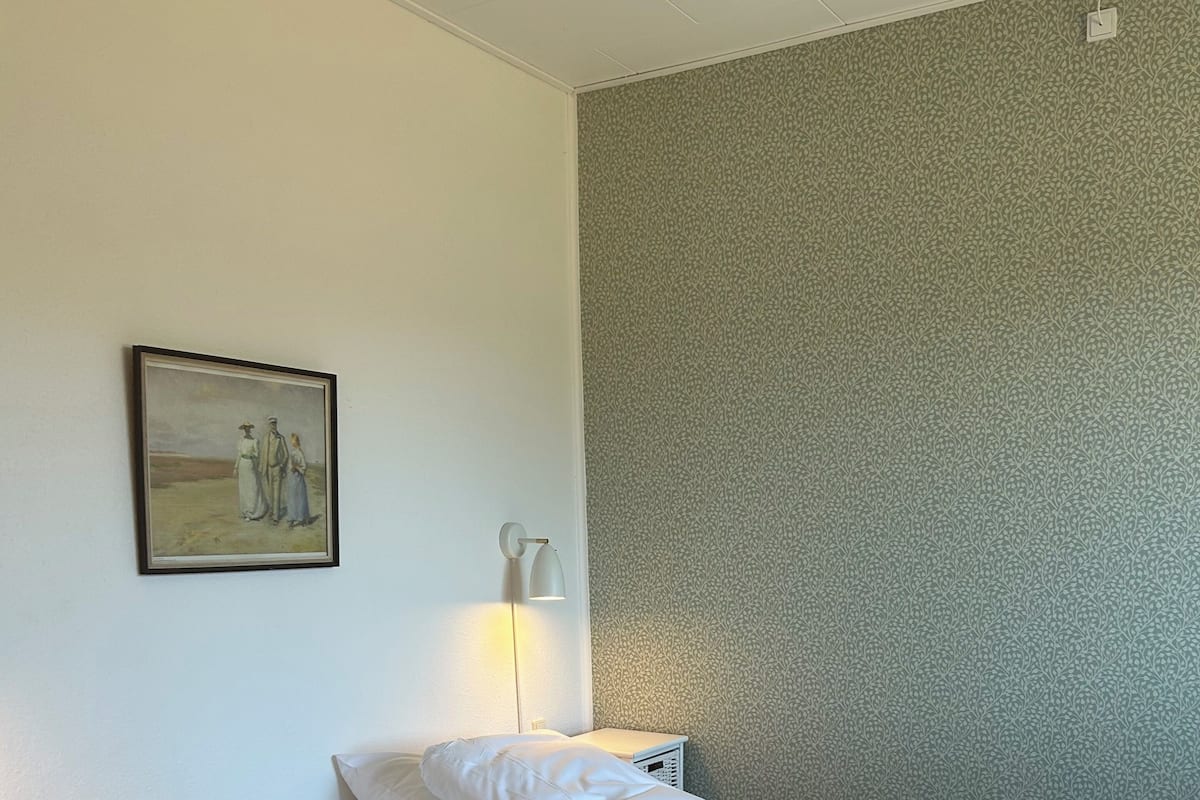 A hotel room with a bed, pillows, a wall-mounted lamp, and a painting.
