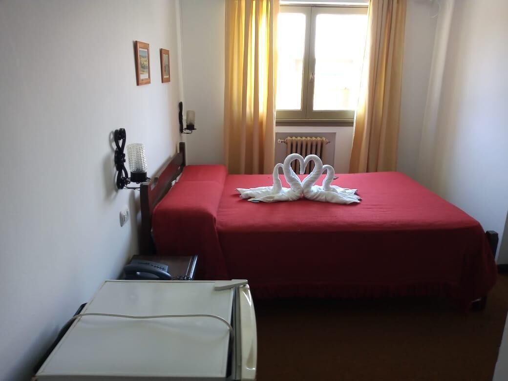 A hotel room with a red bedspread, a white wall-mounted lamp, and a radiator.