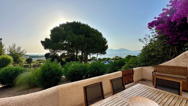 A terrace with a view of the sea, surrounded by greenery and a wooden bench.