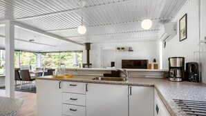 Una cocina moderna con armarios blancos, encimera de granito y lavavajillas integrado. Se ve un comedor con mesa y sillas a través de amplios ventanales.