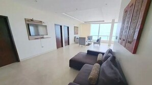 Ein geräumiges Wohnzimmer mit einem großen Flachbildfernseher an der Wand, einem dunkelgrauen Sofa mit Kissen und einem großen Fenster, das für natürliches Licht sorgt.