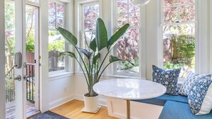 Une pièce lumineuse avec de grandes fenêtres, une plante en pot, une table ronde et des coussins à motifs bleus.